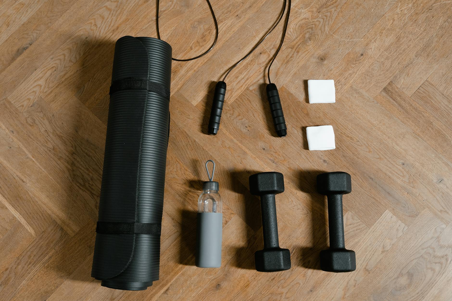 A rolled mat, jump rope, water bottle and two dumbbells laid out on a wooden floor