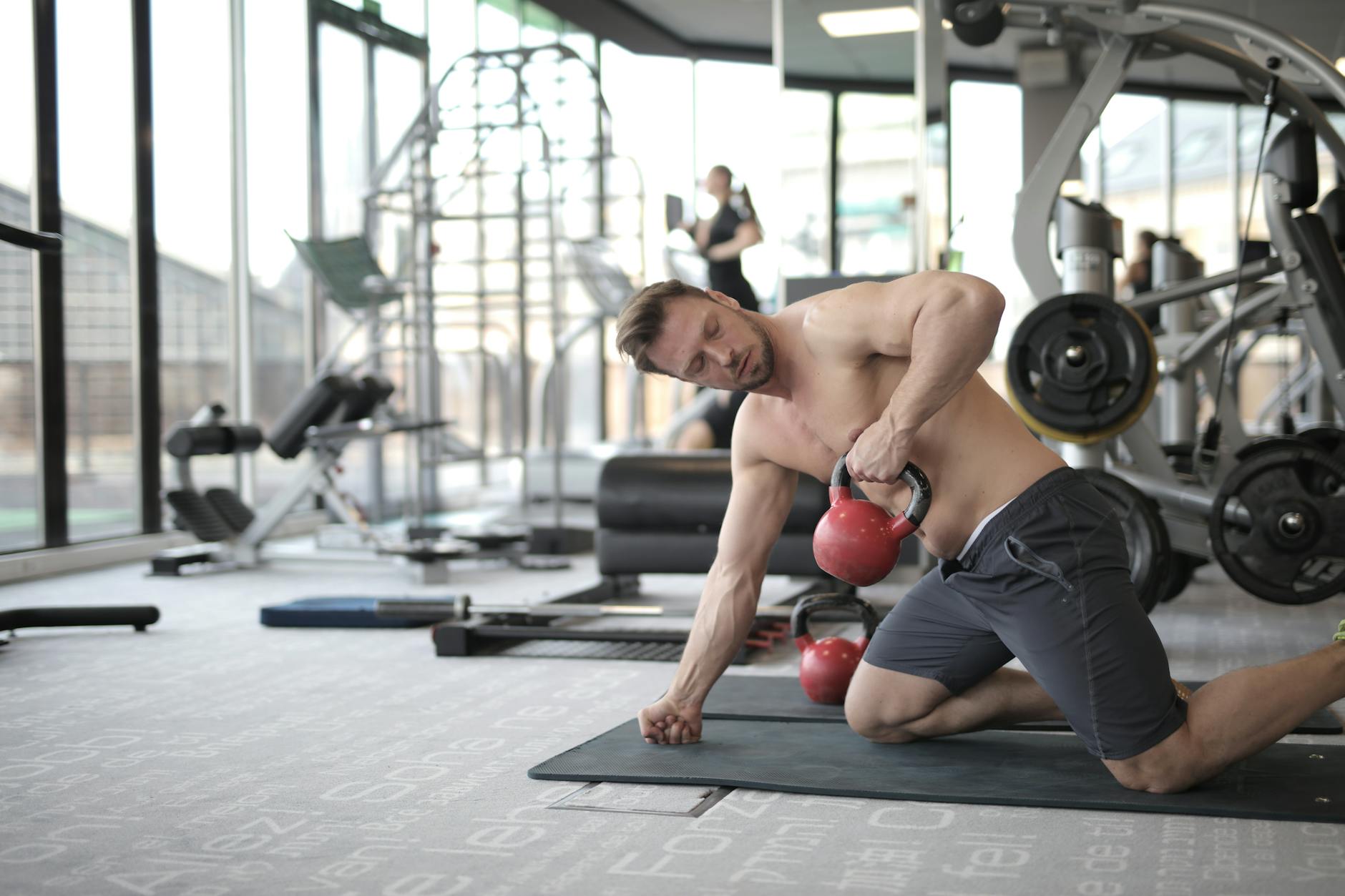 A man doing a kettlebell row on a mat in a bright gym with machines behind him